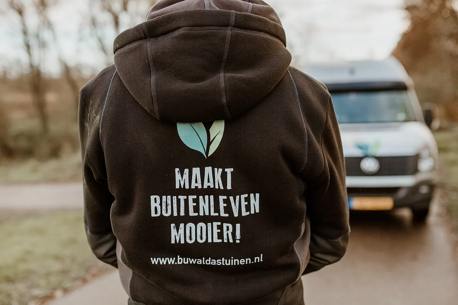Rug van een Buwalda's tuinen-hoodie met de tekst "Maakt buitenleven mooier!", met de bedrijfsbus op de achtergrond
