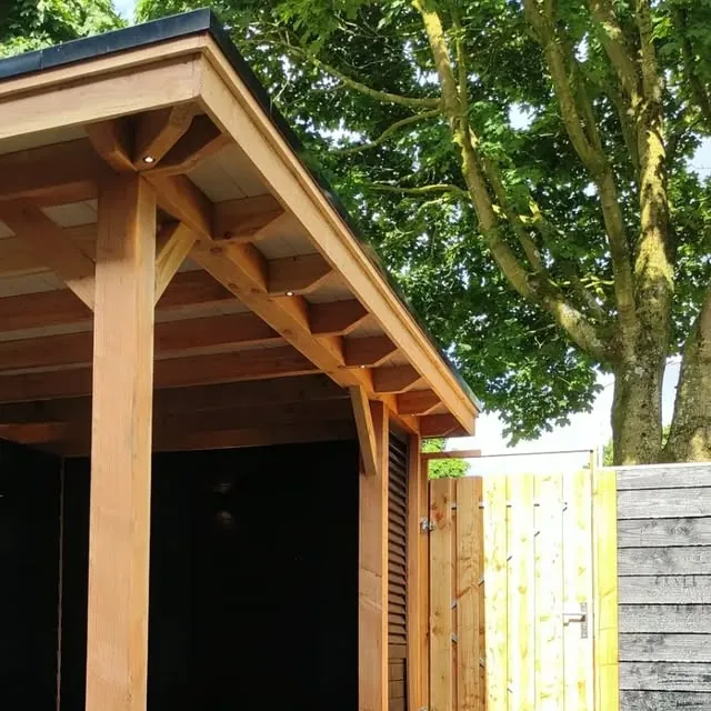 Houten buitenverblijf van lariks met een zadeldak, naast een houten poort onder een grote boom
