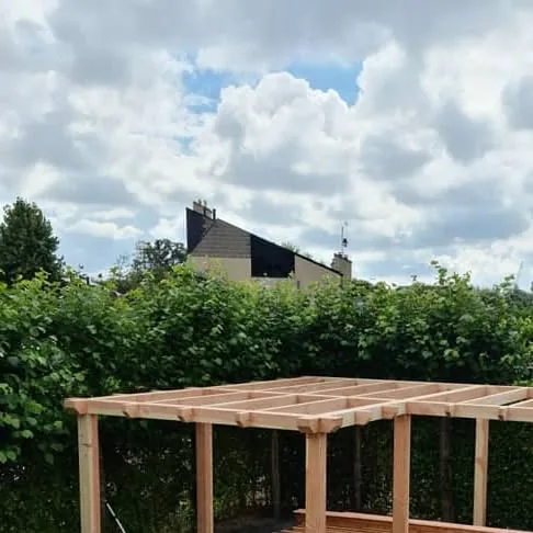Houten geraamte van een hoekmodel buitenverblijf in aanbouw, voor een hoge groene haag