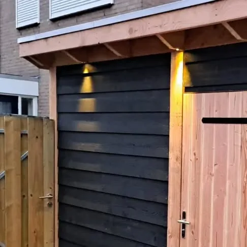 Zwarte houten schuur met lichte houten deur, verlichting onder de overstek en een nieuwe houten poort