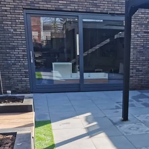 Terras van grote grijze tegels onder een antraciet overkapping, met een plantvak langs de gevel